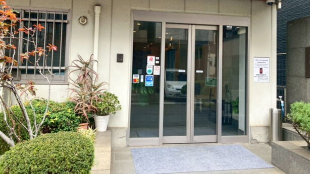 A building entrance with a glass door, a small garden area with potted plants, and a blue mat at the entrance. - HOTEL LiVEMAX BUDGET Kawasaki Daishi (Kawasaki)