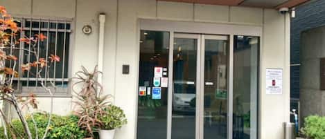 A building entrance with a glass door, a small garden area with potted plants, and a blue mat at the entrance.