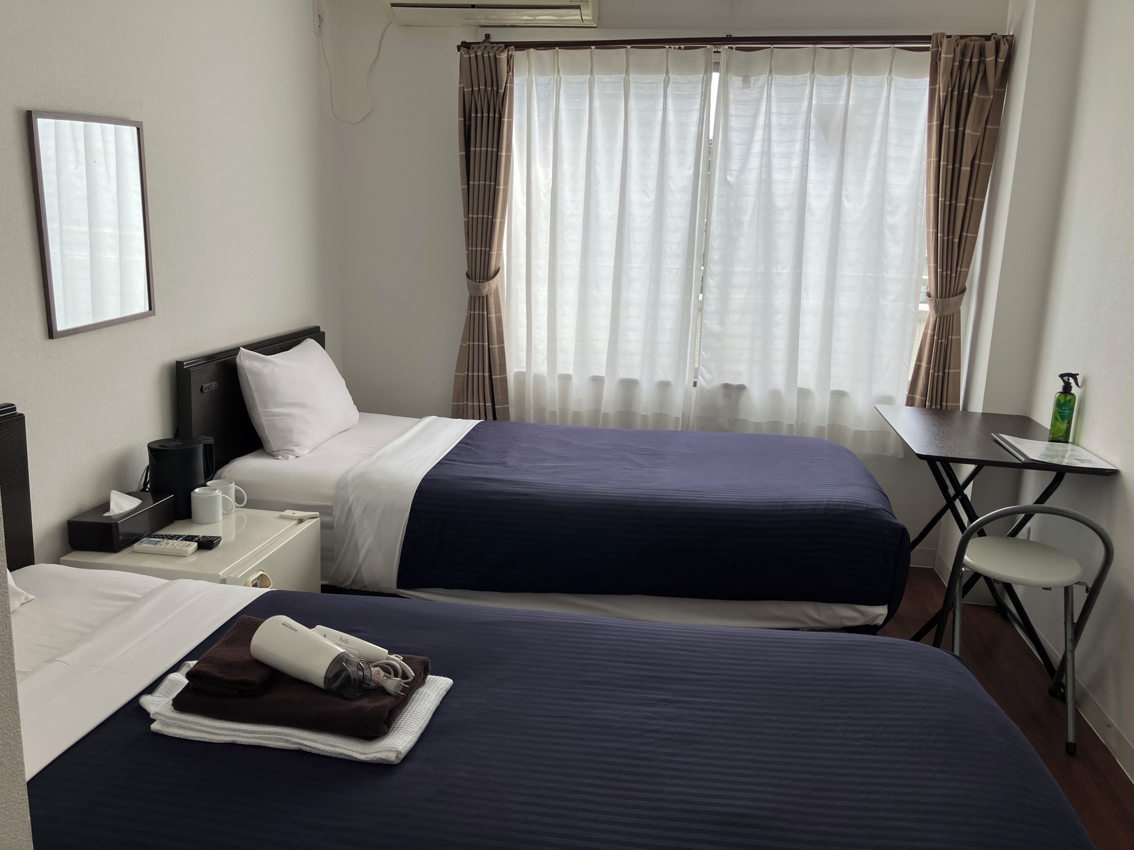 A hotel room with two beds, a bedside table, a chair, and a window with curtains.
