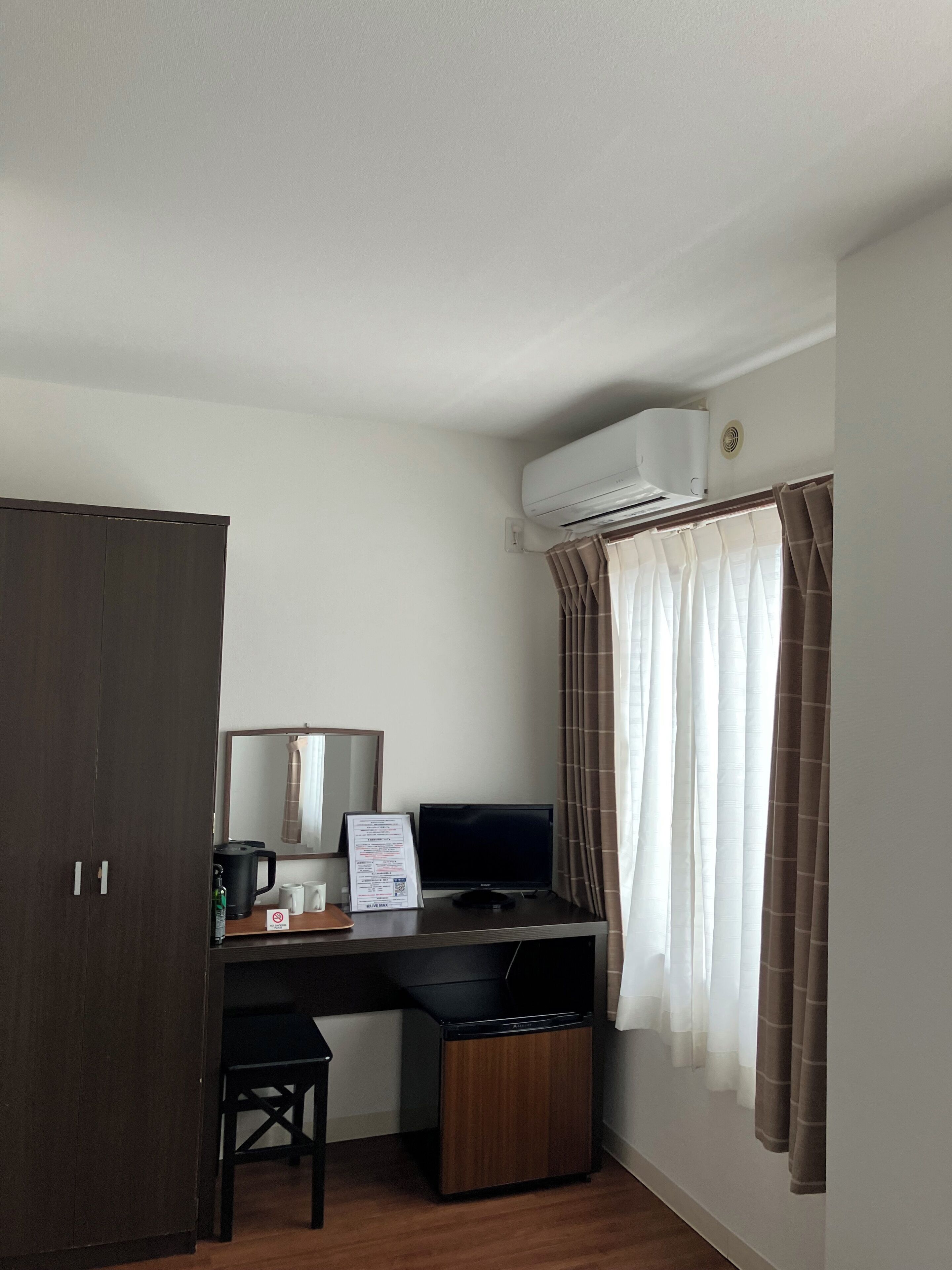 A compact hotel room with a desk, a chair, a small trash bin, a television, and an air conditioning unit.