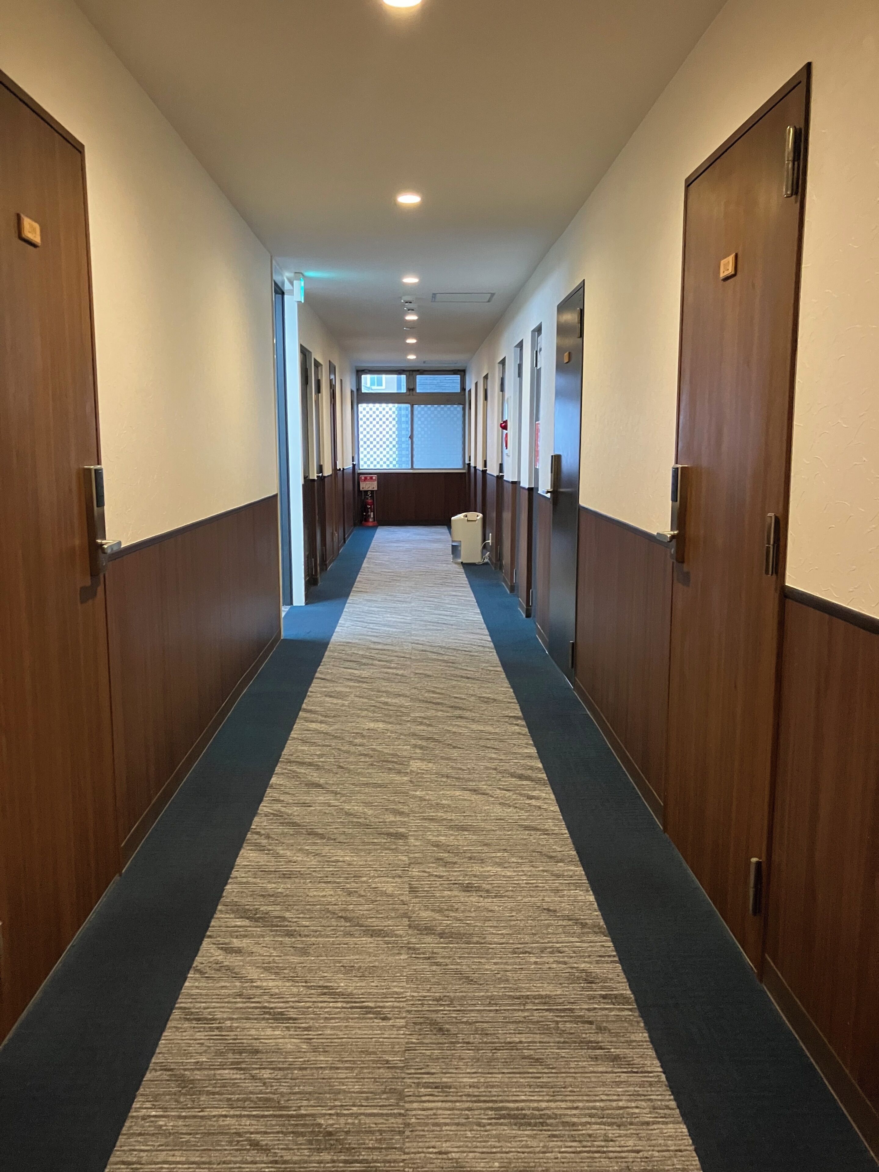 A hotel corridor with wooden doors, a carpeted floor, and a view of the lobby area.