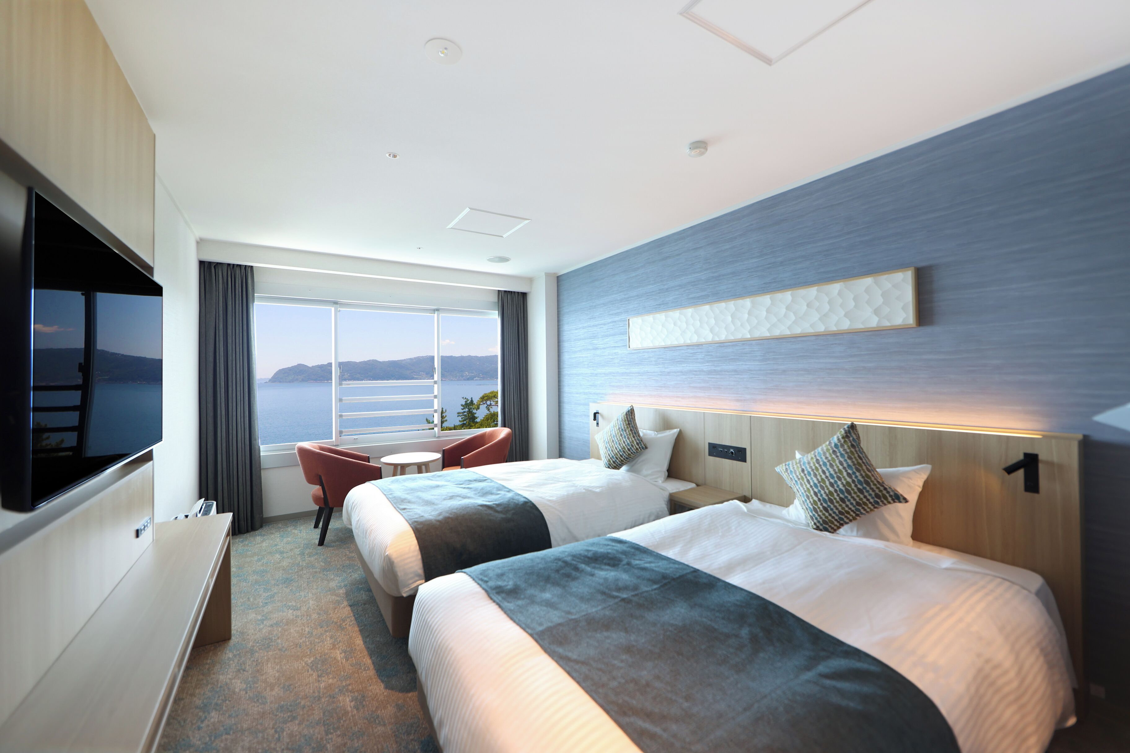 Quarto de hotel com duas camas, televisão, escrivaninha e vista para o mar.
