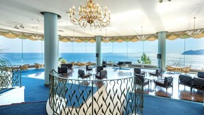 A spacious lounge area with a view of the sea, featuring a chandelier, seating arrangements, and a decorative railing. - Hotel New Akao (Atami)