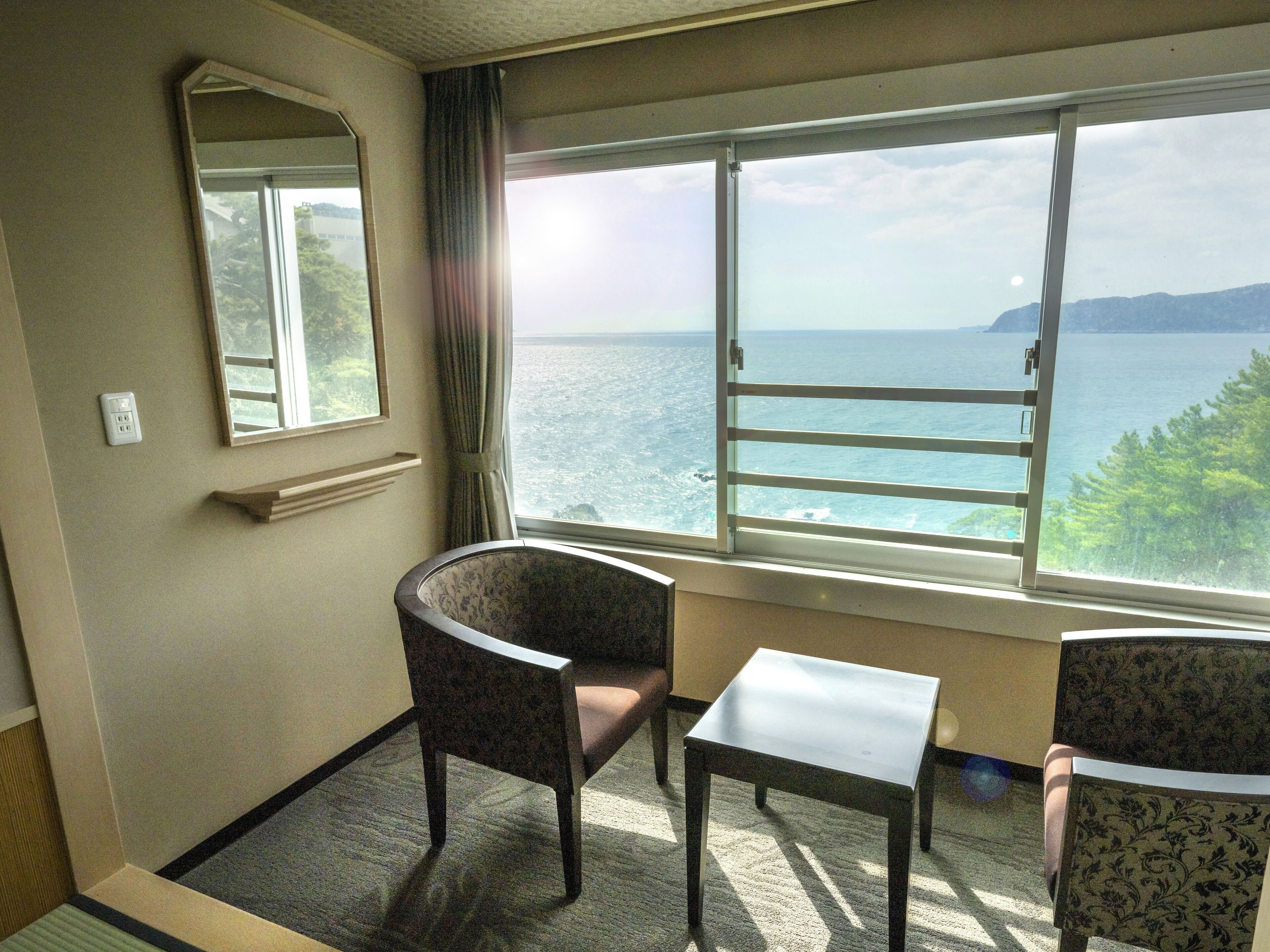A hotel room with a large window offering a view of the sea and a distant island. There is a small table and two chairs.