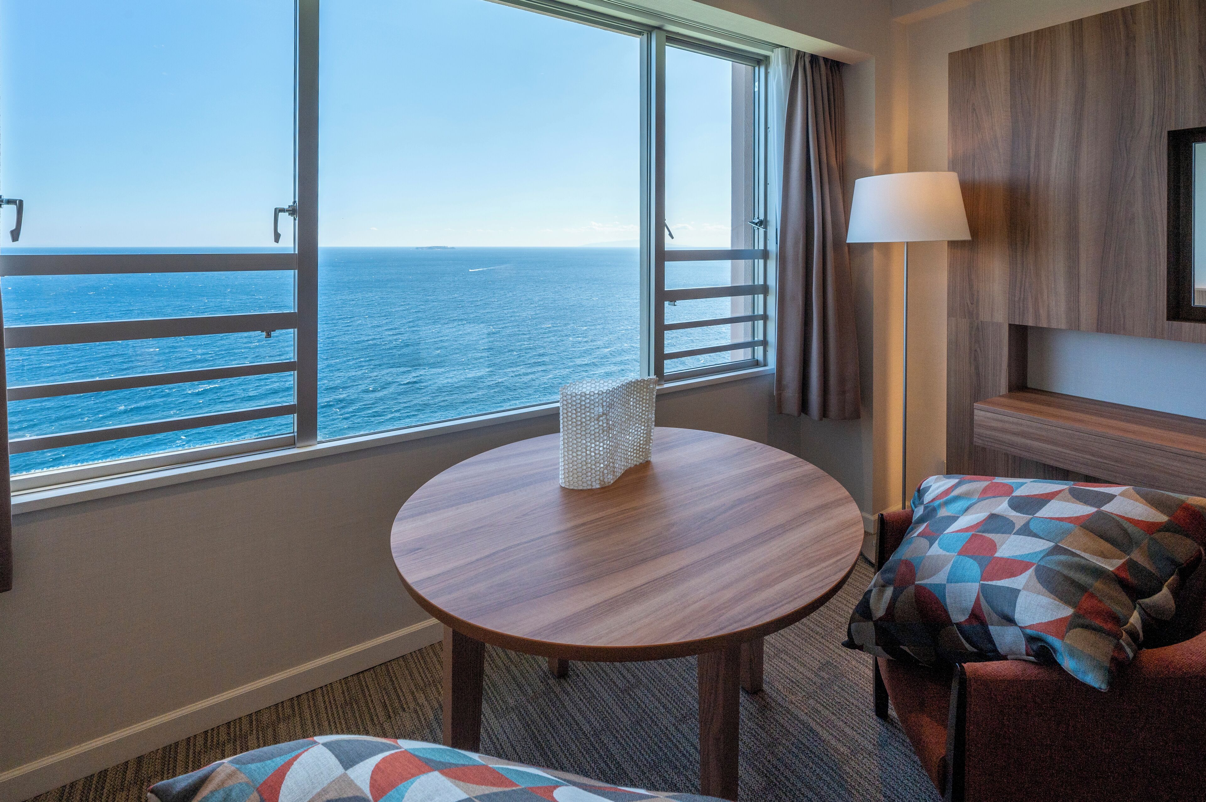 A hotel room with a large window offering a sea view, a round wooden table, a sofa with patterned cushions, and a lamp.