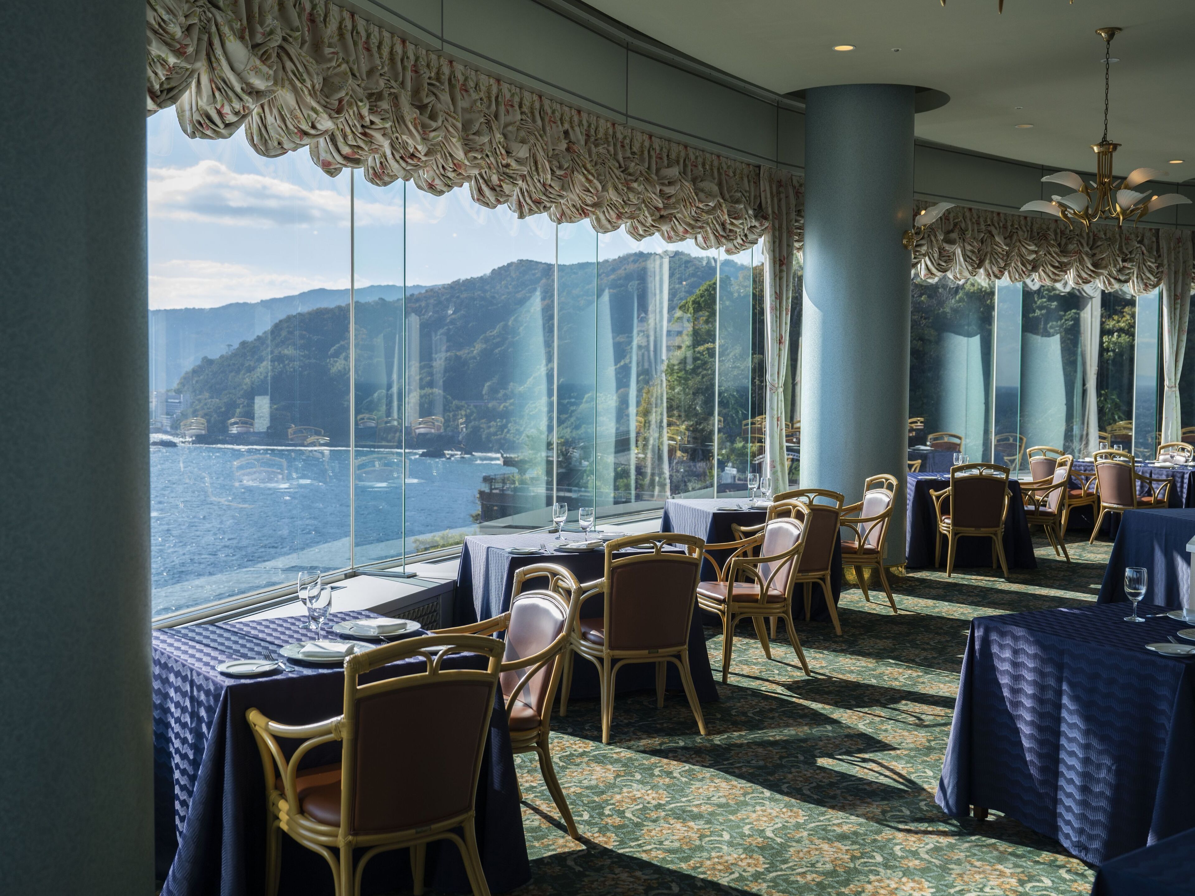 A restaurant with tables set for dining, large windows offering a view of the sea and hills, and a chandelier hanging from the ceiling.