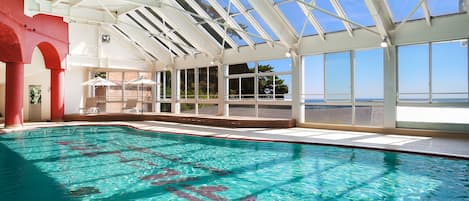 An indoor swimming pool with clear blue water, large windows, and a view of the sea.