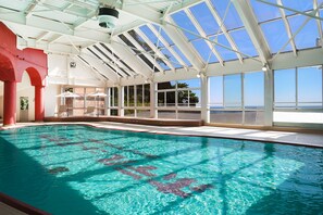 An indoor swimming pool with clear blue water, large windows, and a view of the sea.