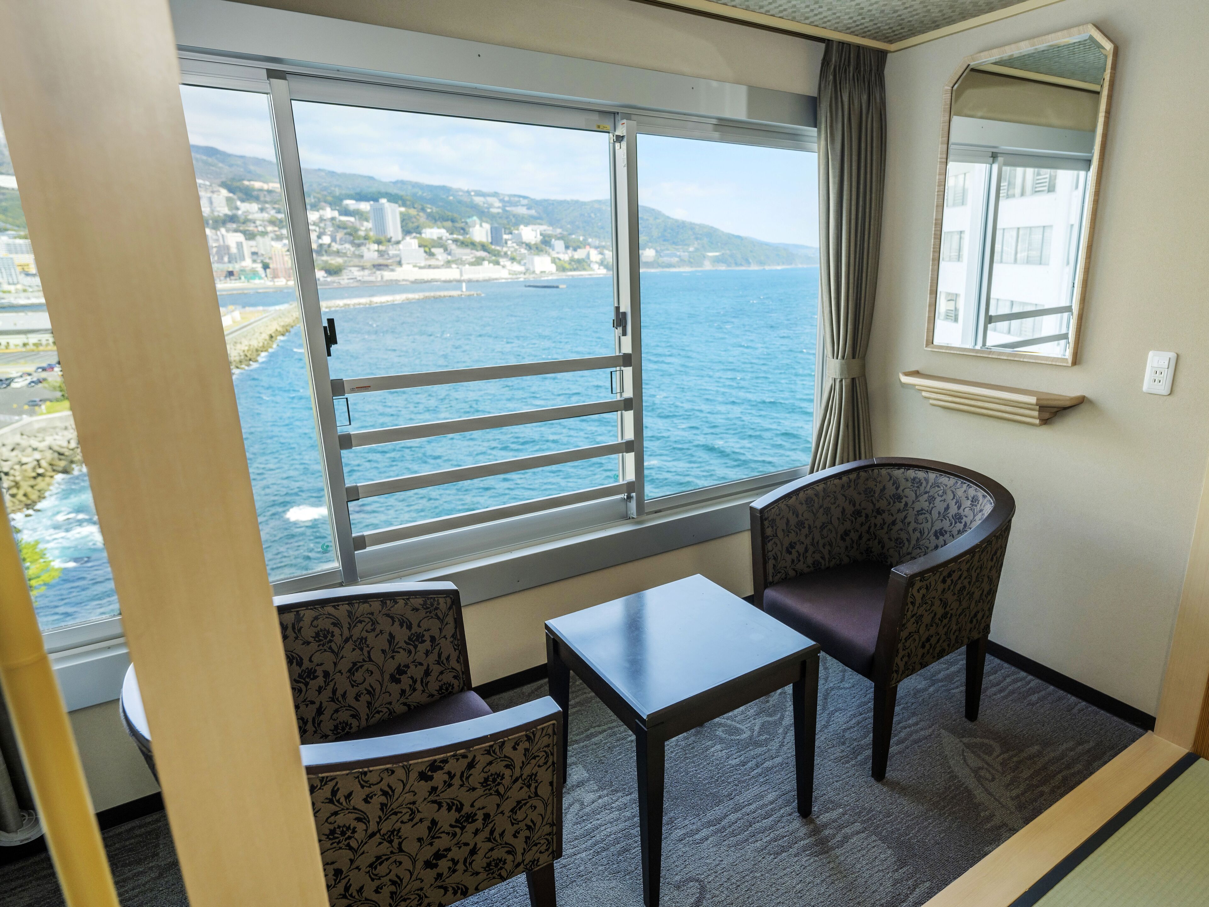 A room with a large window offering a view of the sea and a cityscape. There is a small table and two patterned chairs.