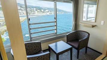A room with a large window offering a view of the sea and a cityscape. There is a small table and two patterned chairs.