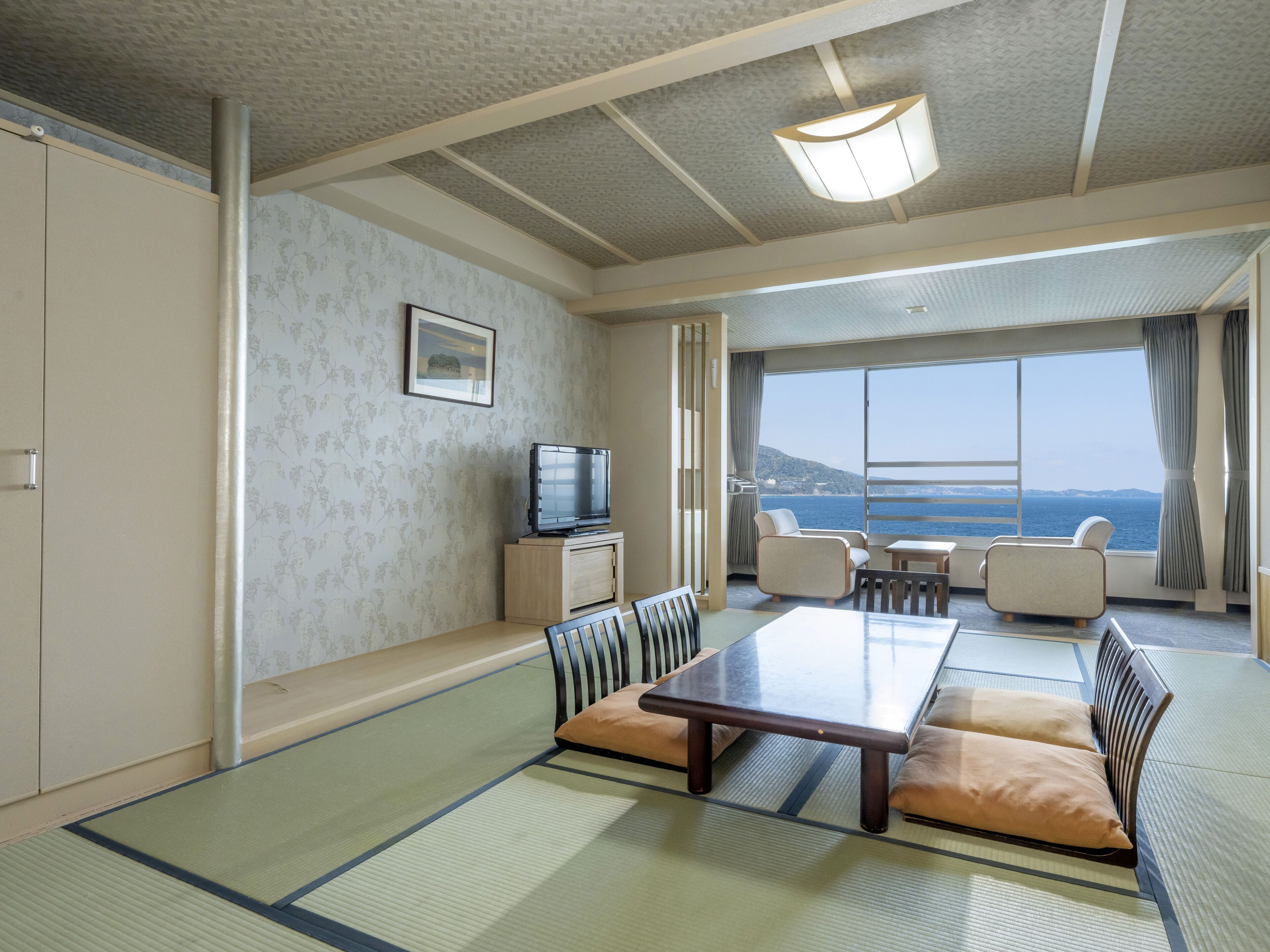 Una habitación tradicional japonesa con suelo de tatami, una mesa baja y asientos. Un televisor está montado en un mueble, y la habitación tiene ventanas amplias con vista al mar.