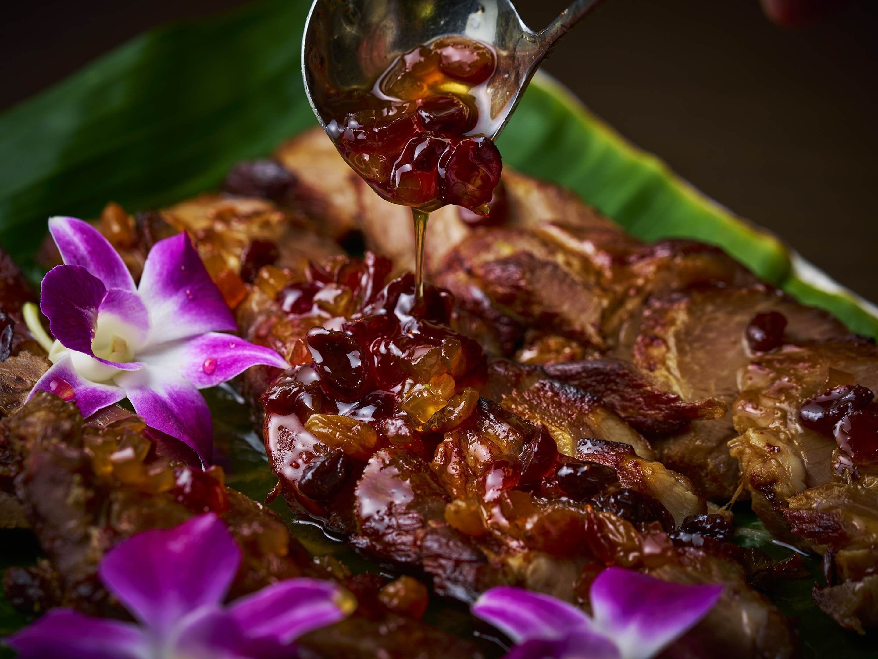 A close-up of a dish with meat and a sauce being poured over it, accompanied by purple flowers.