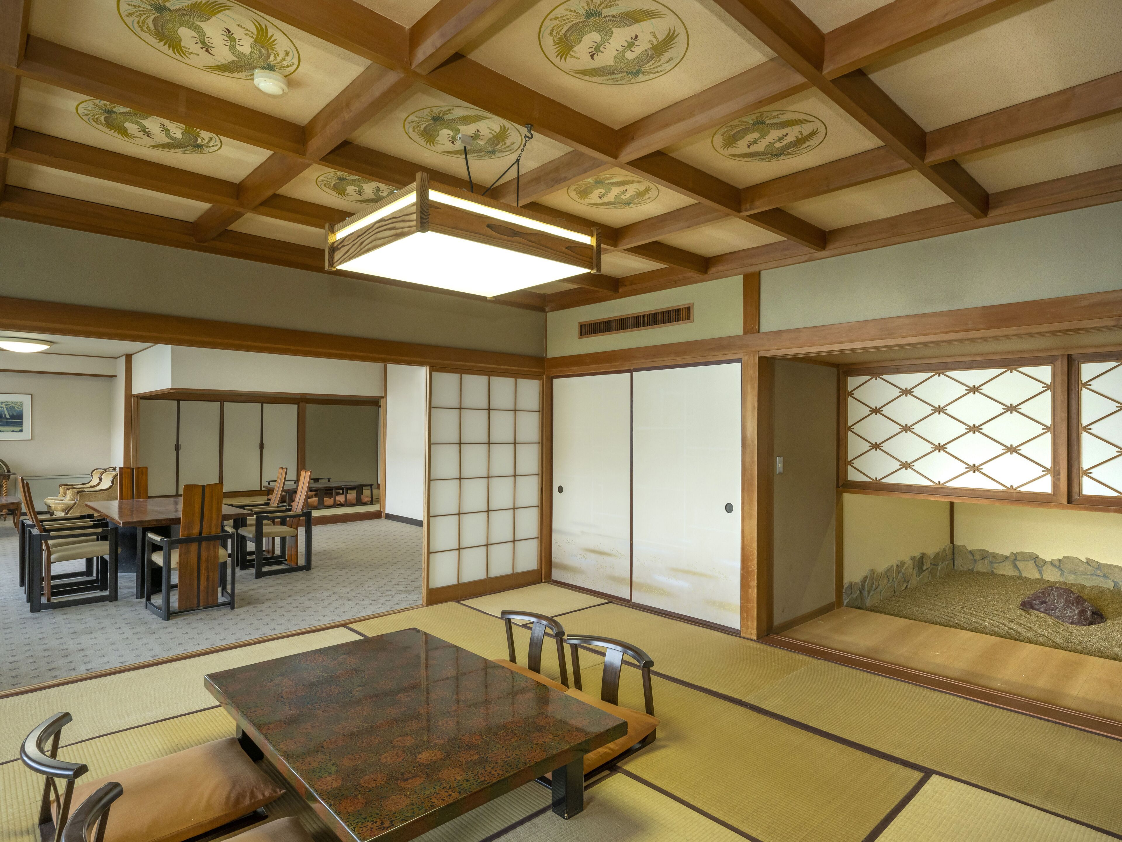A traditional Japanese room with tatami flooring, a low table with chairs, sliding doors, and a ceiling with wooden beams and decorative patterns.
