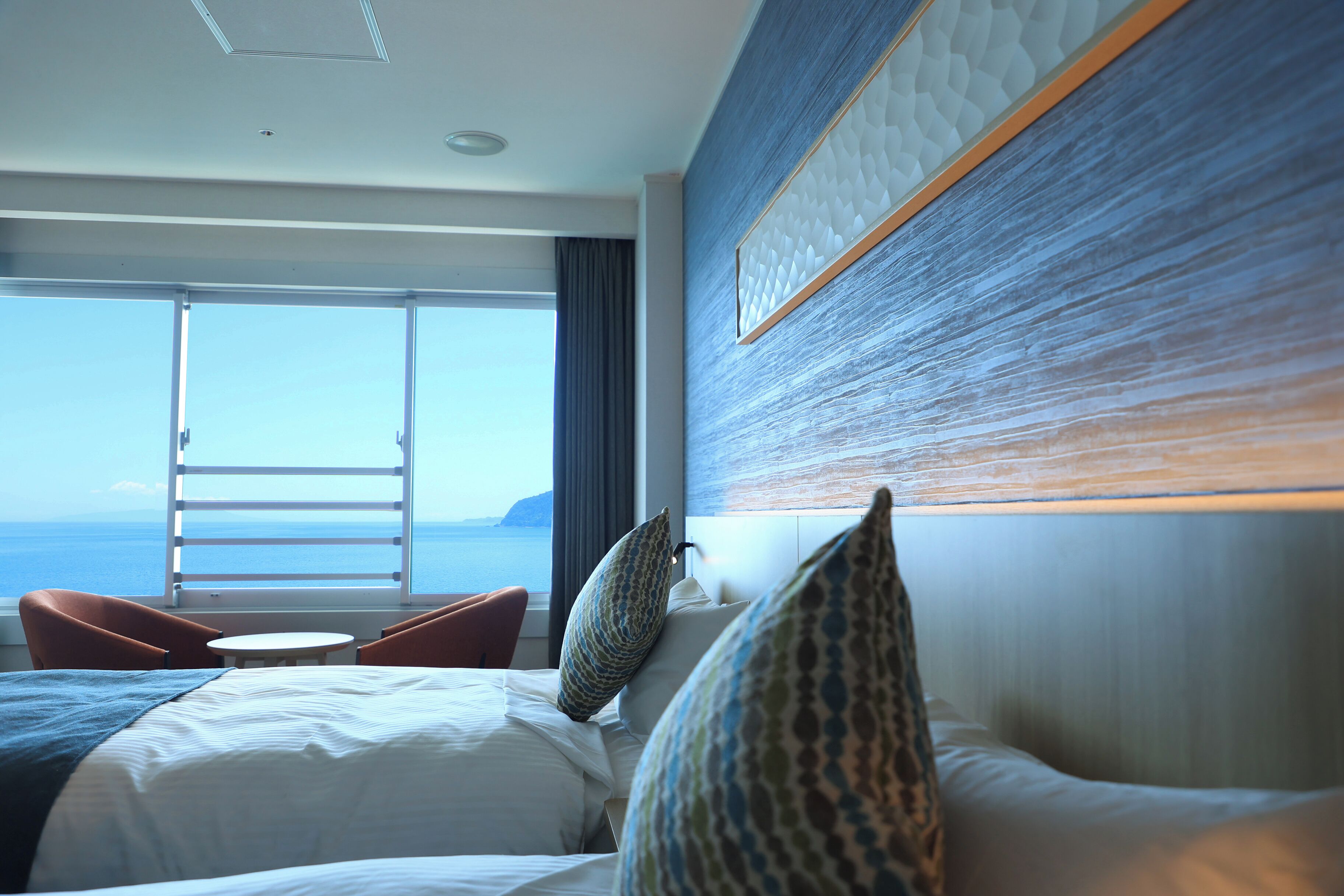 A hotel room with a bed, two chairs, a small table, and a large window offering a view of the sea.