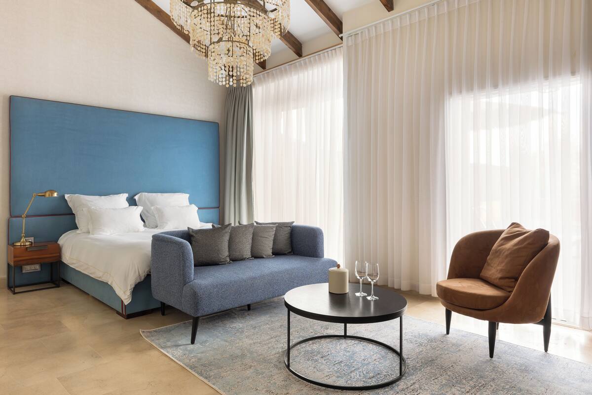 Une chambre à coucher moderne avec une tête de lit bleue, un canapé gris, un fauteuil marron, une table basse ronde et un lustre.