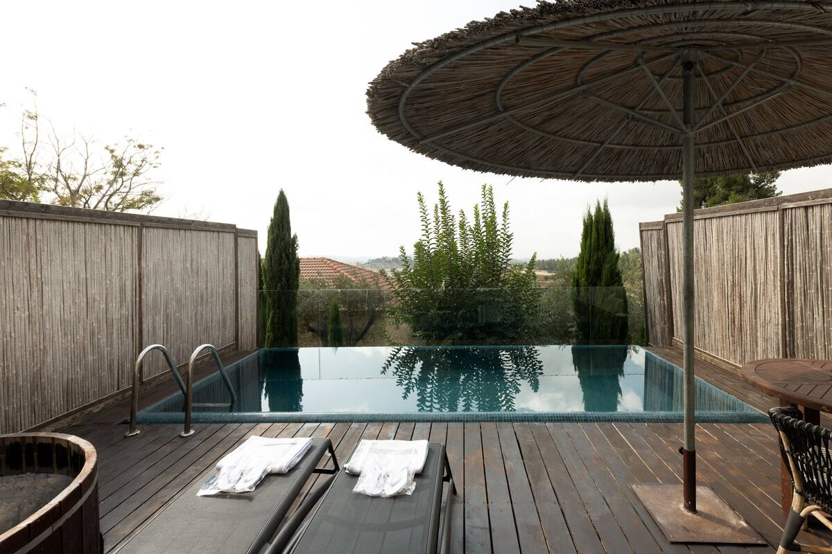 Une terrasse en bois avec une piscine à débordement en verre, des chaises longues et un parasol en chaume.