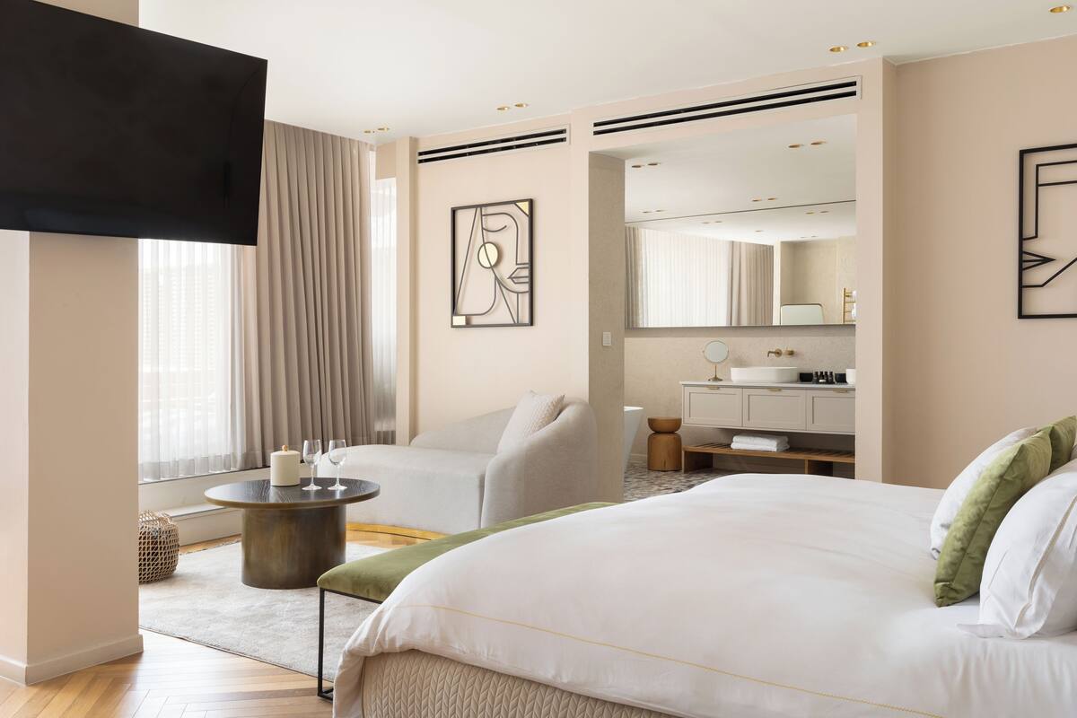 Une chambre d’hôtel moderne avec un grand lit, une table basse ronde, un canapé, une télévision et une salle de bain visible à travers une porte ouverte.