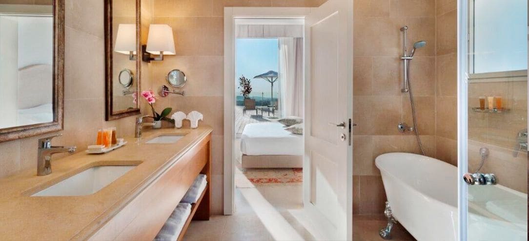 A modern bathroom with a double sink, a bathtub, and a shower. There is a visible pathway leading to a bedroom with a bed and a view of the outdoors.