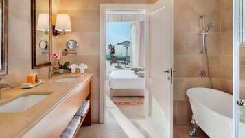 A modern bathroom with a double sink, a bathtub, and a shower. There is a visible pathway leading to a bedroom with a bed and a view of the outdoors.