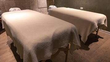 Zwei Massagetische mit weißen Laken in einem Raum mit Steinwänden und Holzboden.