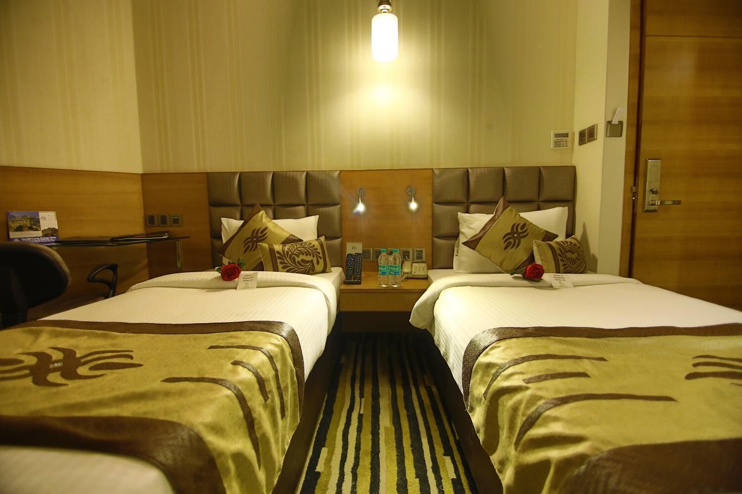 A hotel room with two beds, a desk, a chair, a nightstand with a phone and a lamp, and a door.