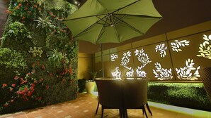 A modern outdoor seating area with a green umbrella, a vertical garden wall, and illuminated leaf patterns on the wall. - JRD Exotica (New Delhi)