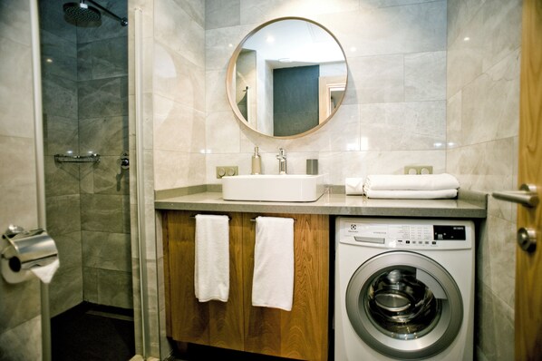 A modern bathroom with a shower, a sink, a mirror, a washing machine, and wooden cabinets. - Degirmenburnu Residence (Bodrum)
