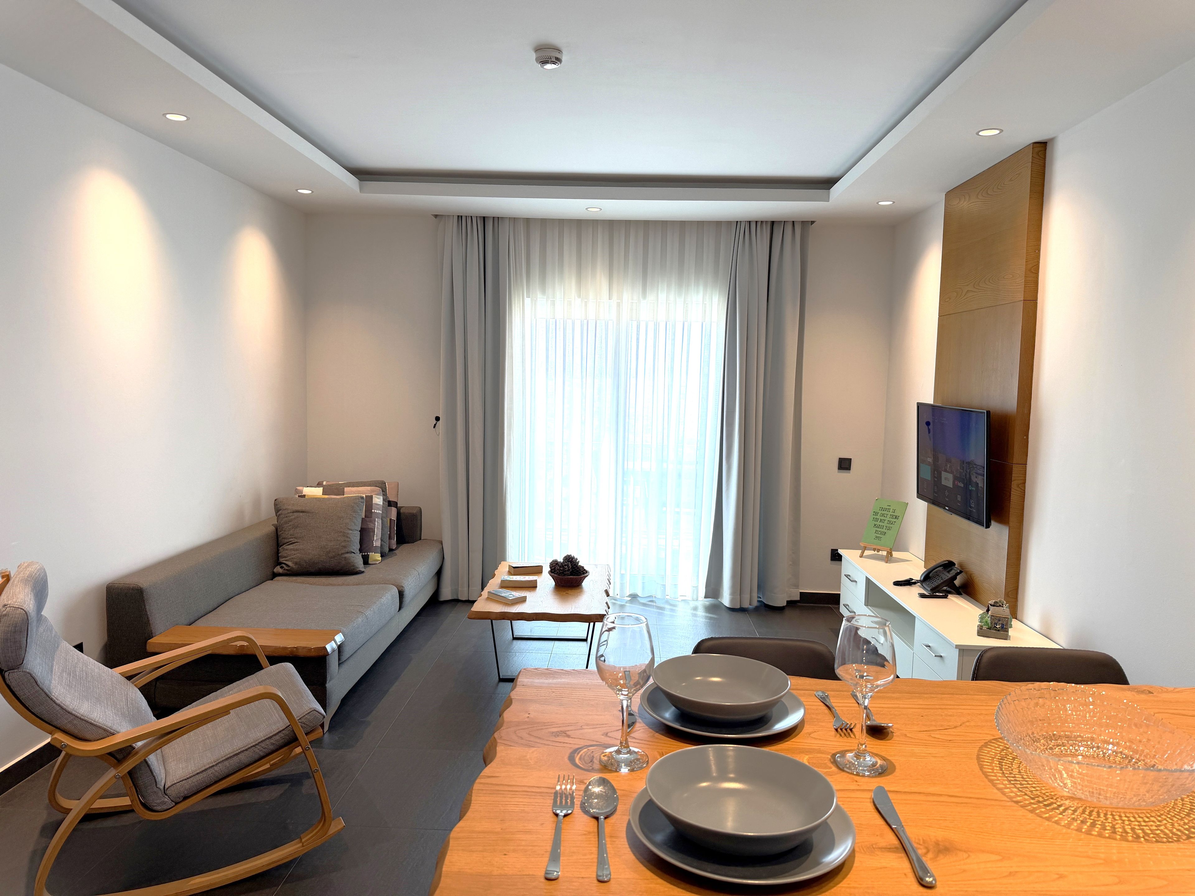 Een moderne hotelkamer met een bank, een eettafel gedekt voor twee personen, een aan de muur bevestigde televisie en een raam met gordijnen.