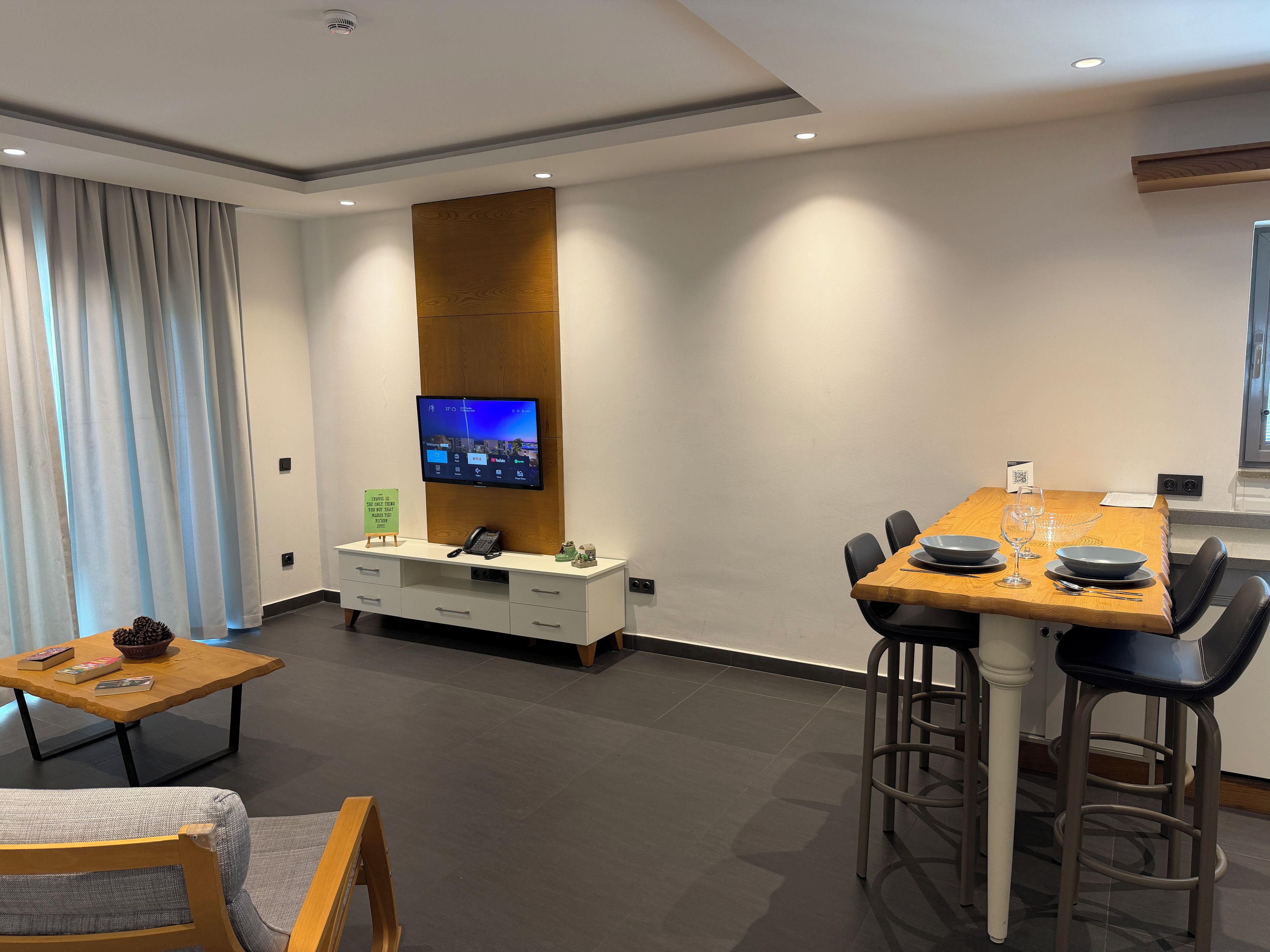 Una habitación de hotel moderna con comedor, un televisor montado en un panel de madera, un escritorio con teléfono y una zona de estar con sofá y sillas.