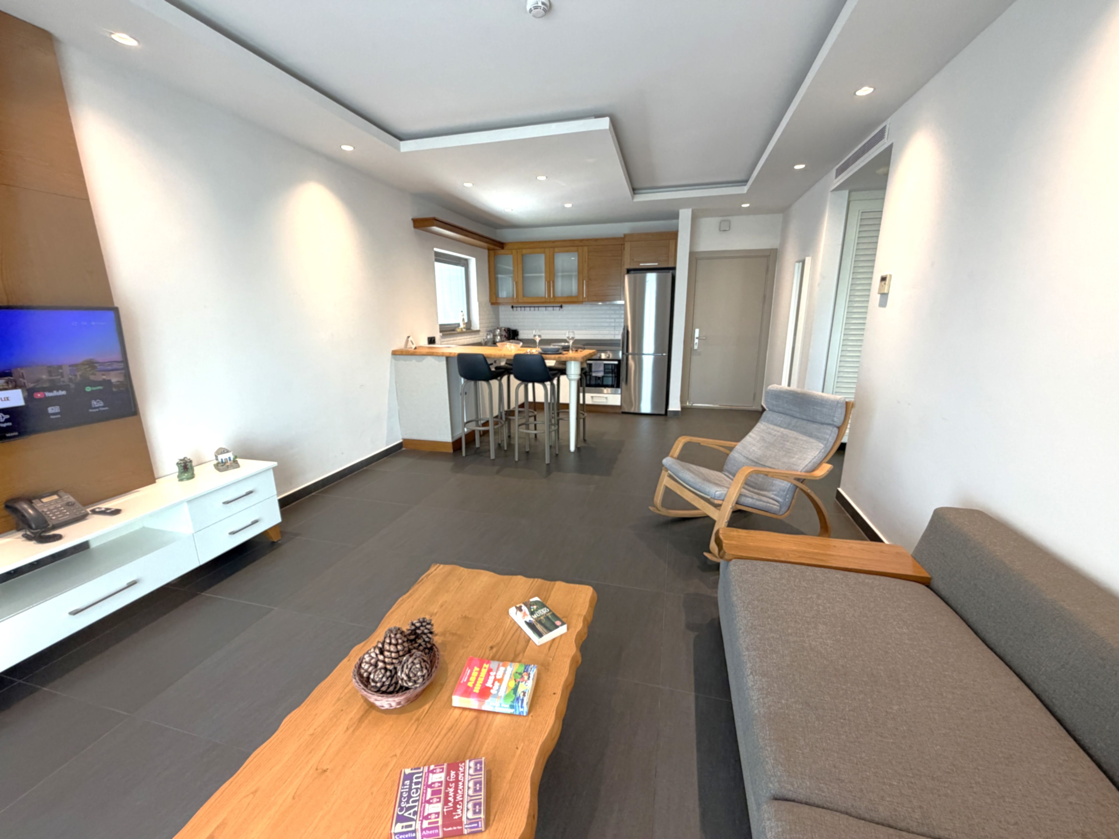 A modern living room with a sofa, a coffee table, and a TV. There is a kitchen in the background with cabinets and appliances.