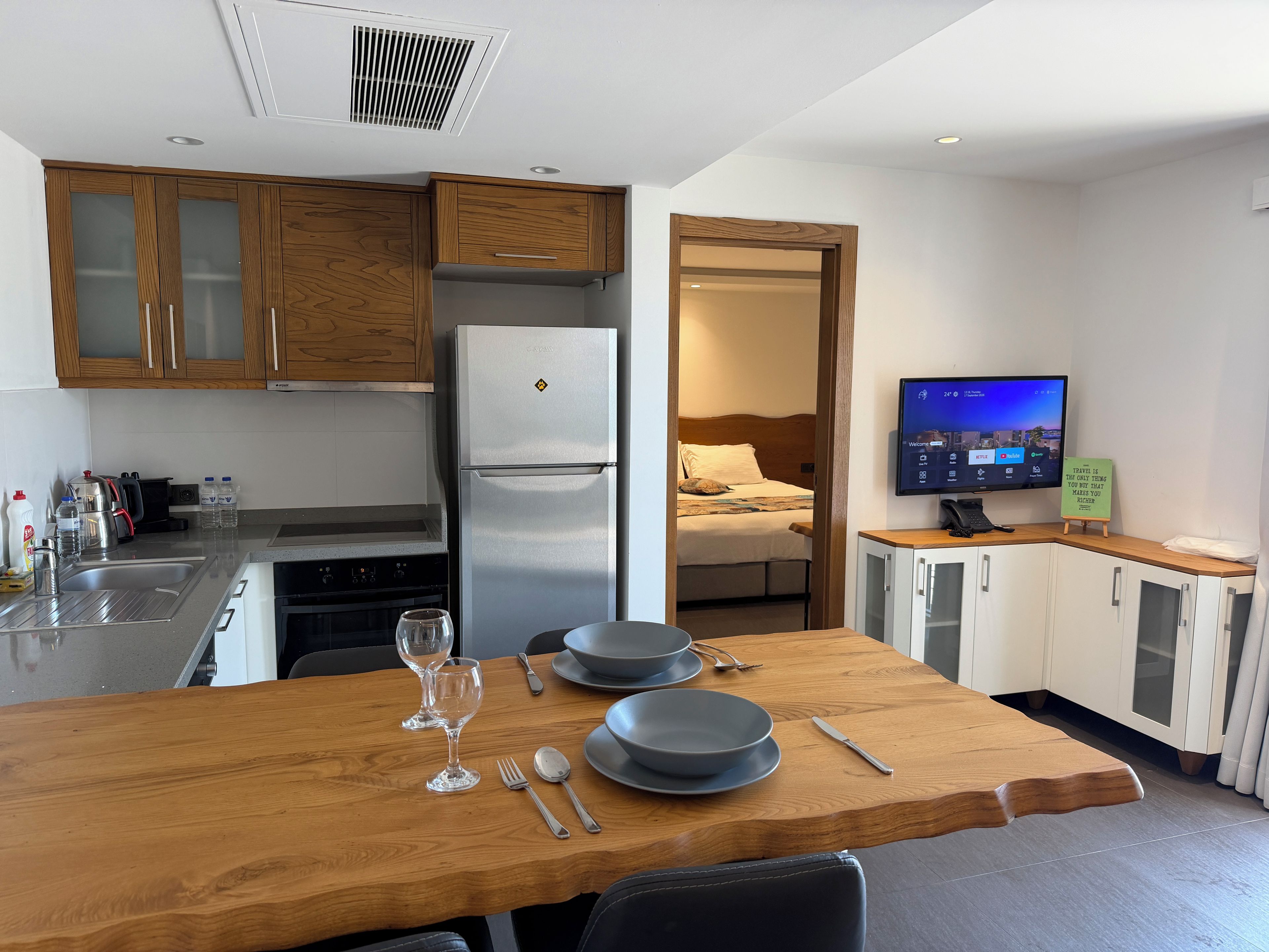 A compact kitchen with a refrigerator, stove, and wooden cabinets. A dining table with plates, cutlery, and glasses is set. A television is mounted on the wall, and a bed is visible through an open door.