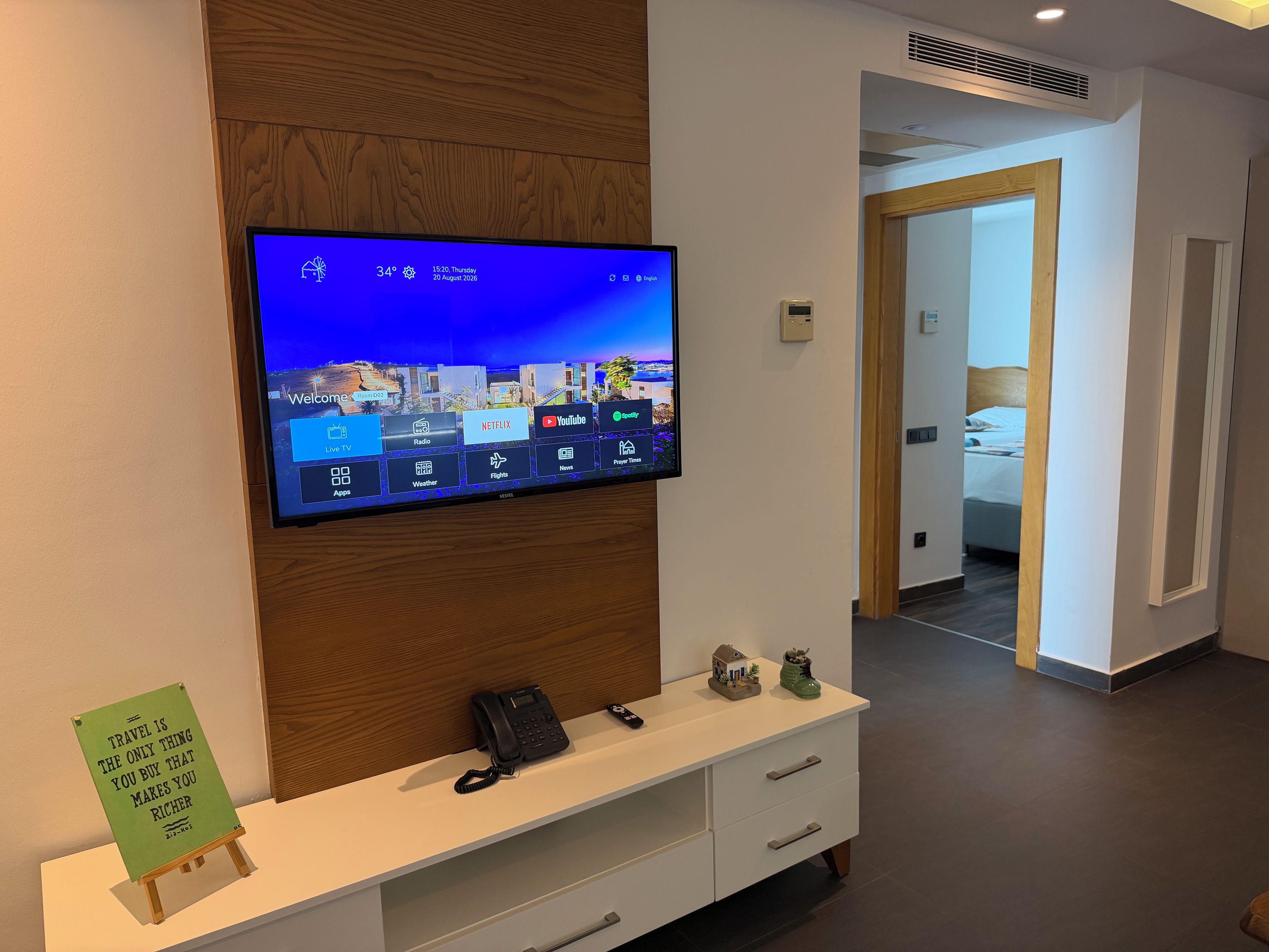 Et hotelreceptionsområde med et ophængt fjernsyn, der viser vejrudsigten, en reception med telefon og et lille skilt, samt udsigt til et soveværelse.