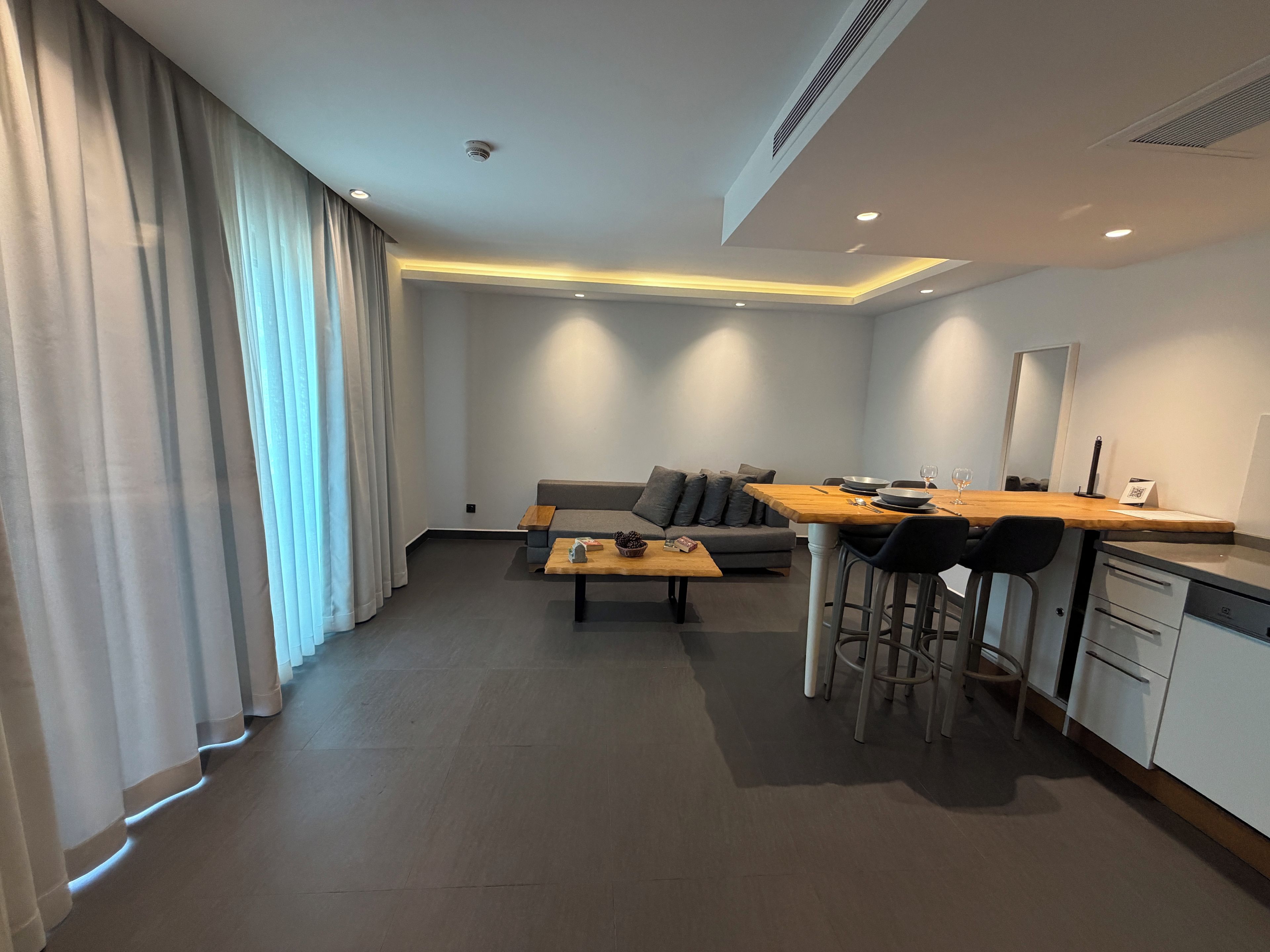 A modern living room with a sofa, a wooden dining table, and a kitchen area with a refrigerator and a sink.
