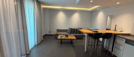 A modern living room with a sofa, a wooden dining table, and a kitchen area with a refrigerator and a sink.