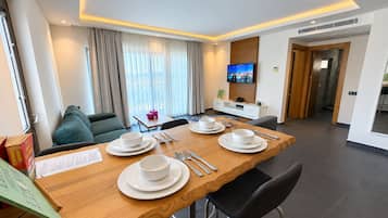 Habitación de hotel moderna con zona de comedor, sofá, televisión y vistas al exterior.