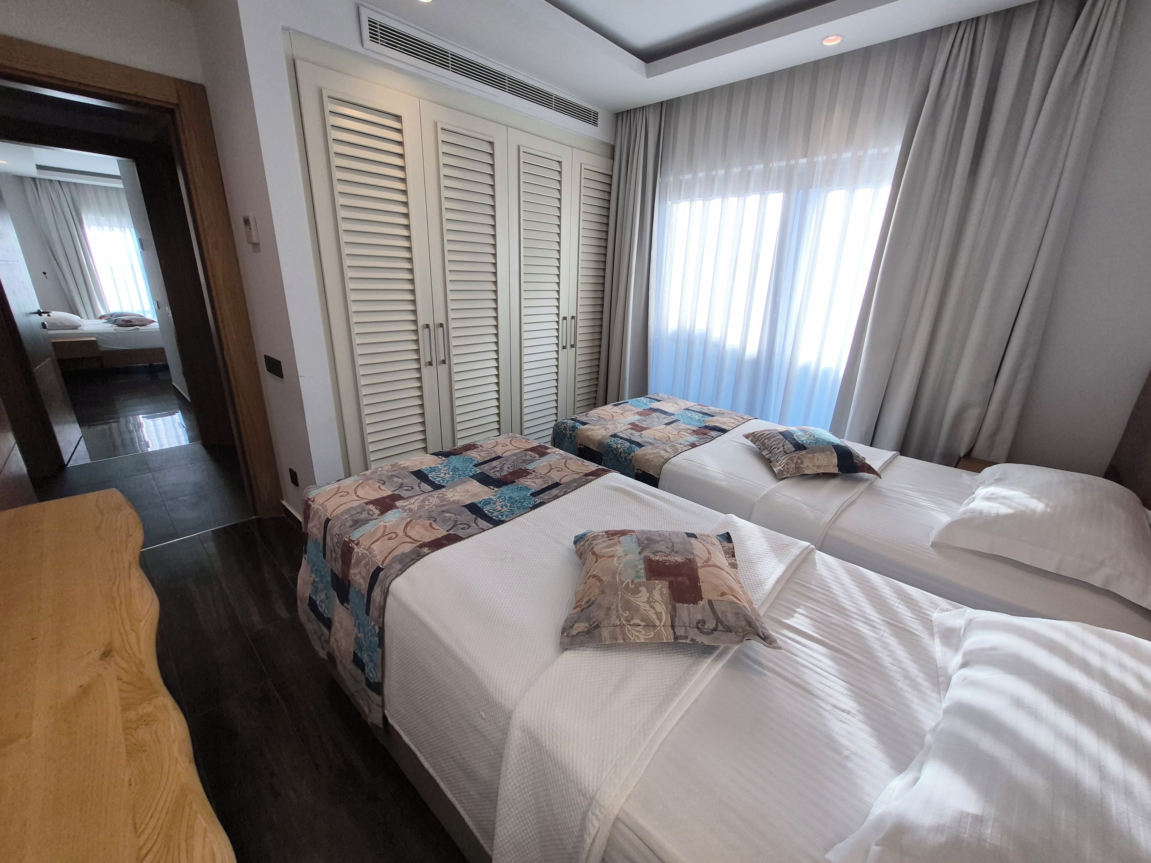 Habitación de hotel con dos camas, un escritorio de madera, un armario con puertas batientes y un amplio ventanal con cortinas.