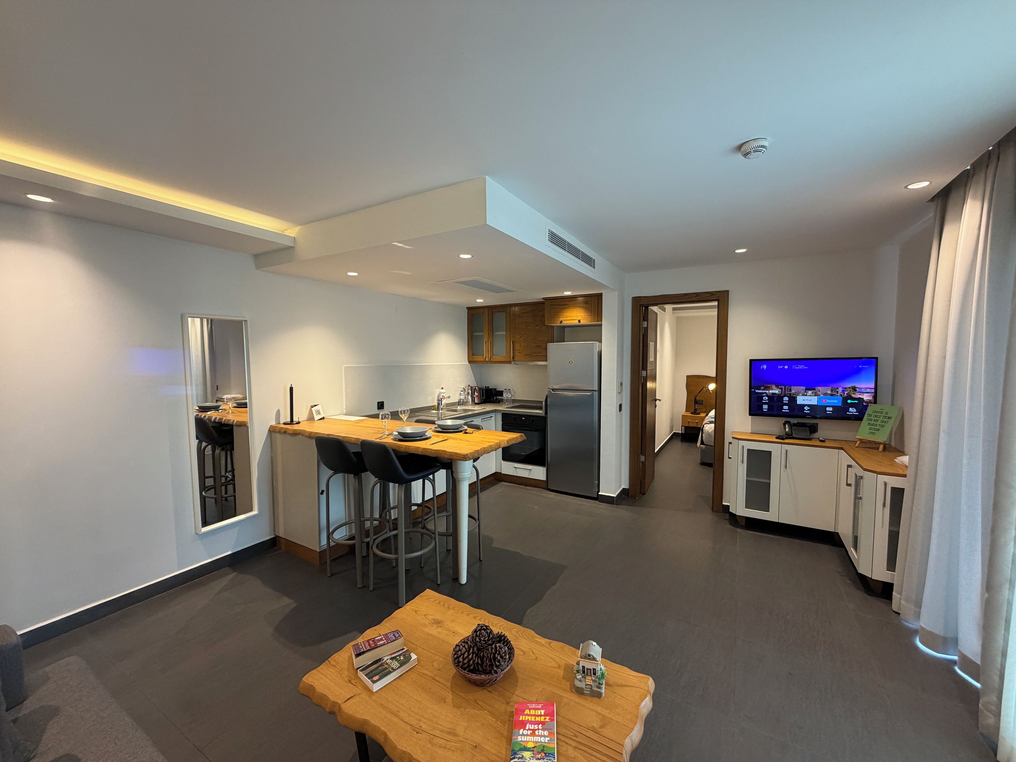 A modern kitchen with a dining area, a small living space with a TV, and a wooden table with books and a pine cone.