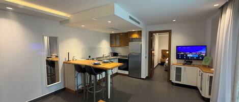 A modern kitchen with a dining area, a small living space with a TV, and a wooden table with books and a pine cone.