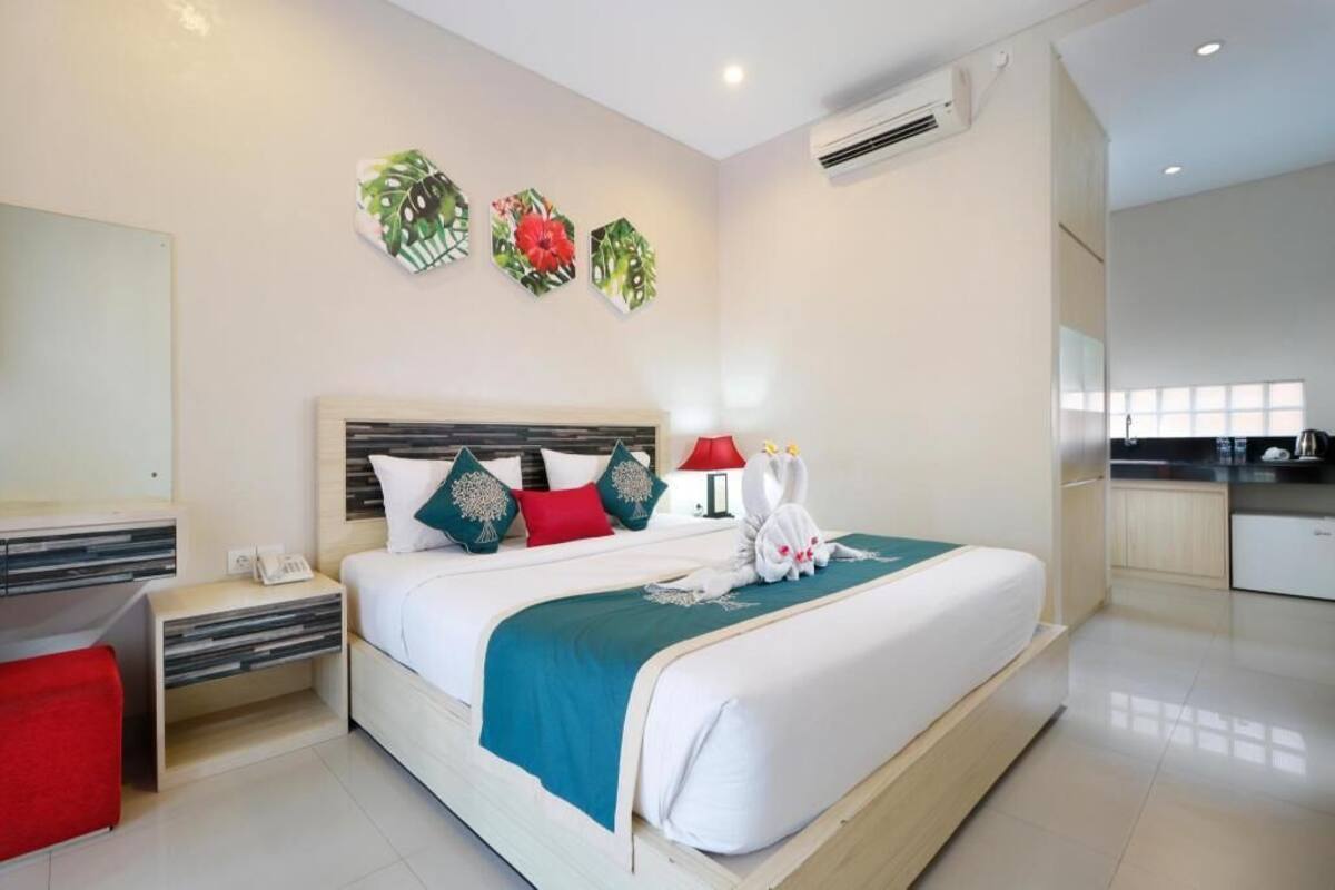A modern hotel room with a bed, bedside tables, a red lamp, and a kitchen area.