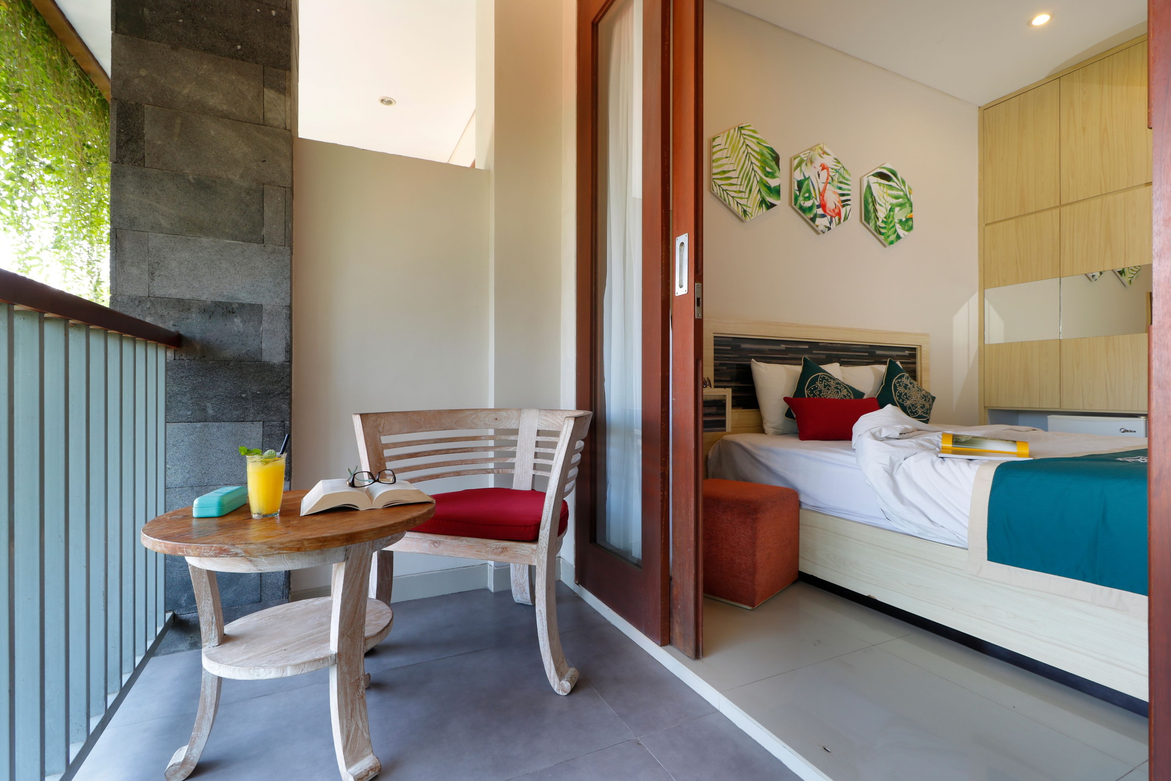 Une chambre avec un lit, une chaise en bois, une petite table avec une boisson, et une vue sur un balcon doté d’un mur en pierre et de verdure.