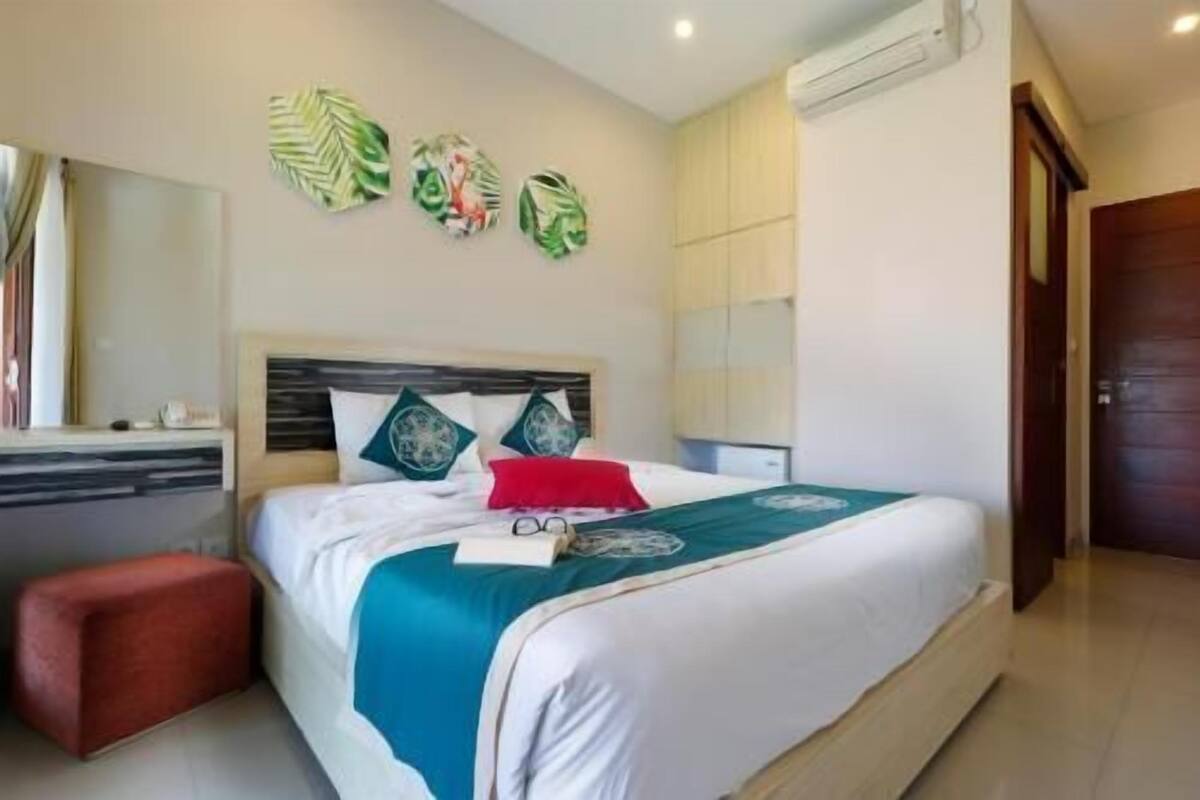 A hotel room with a bed, a desk, a red ottoman, and wall art.