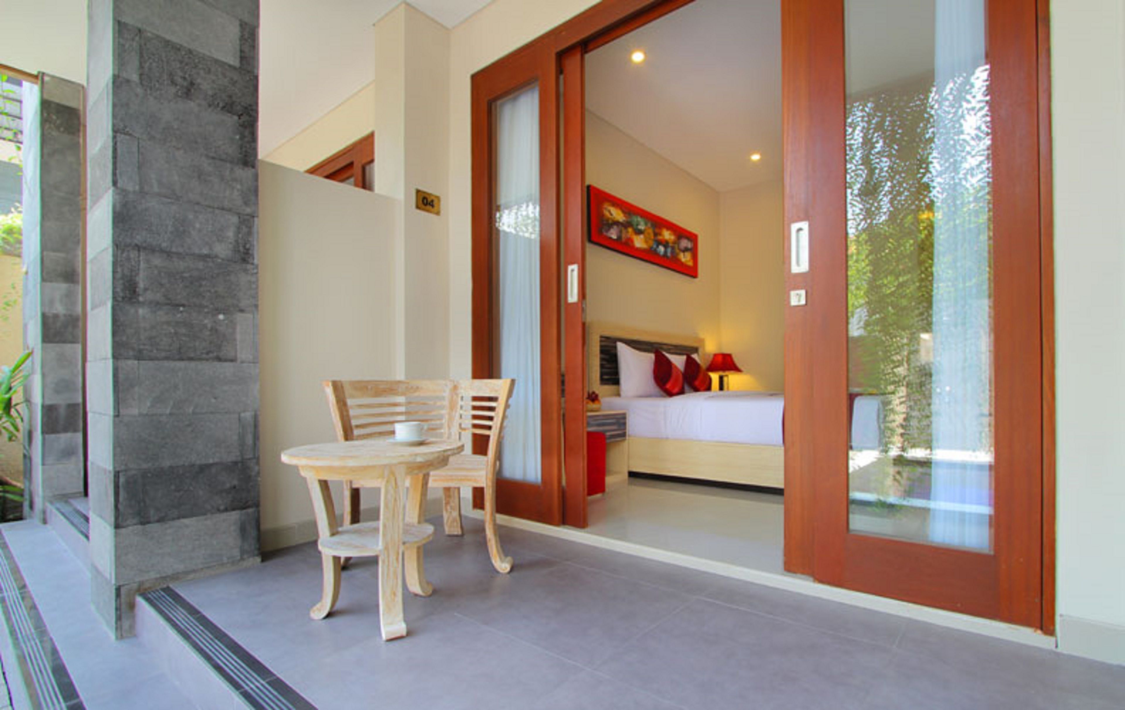 A modern hotel room with a bed, a nightstand, and a red lamp. There is a wooden table and chairs outside the room.