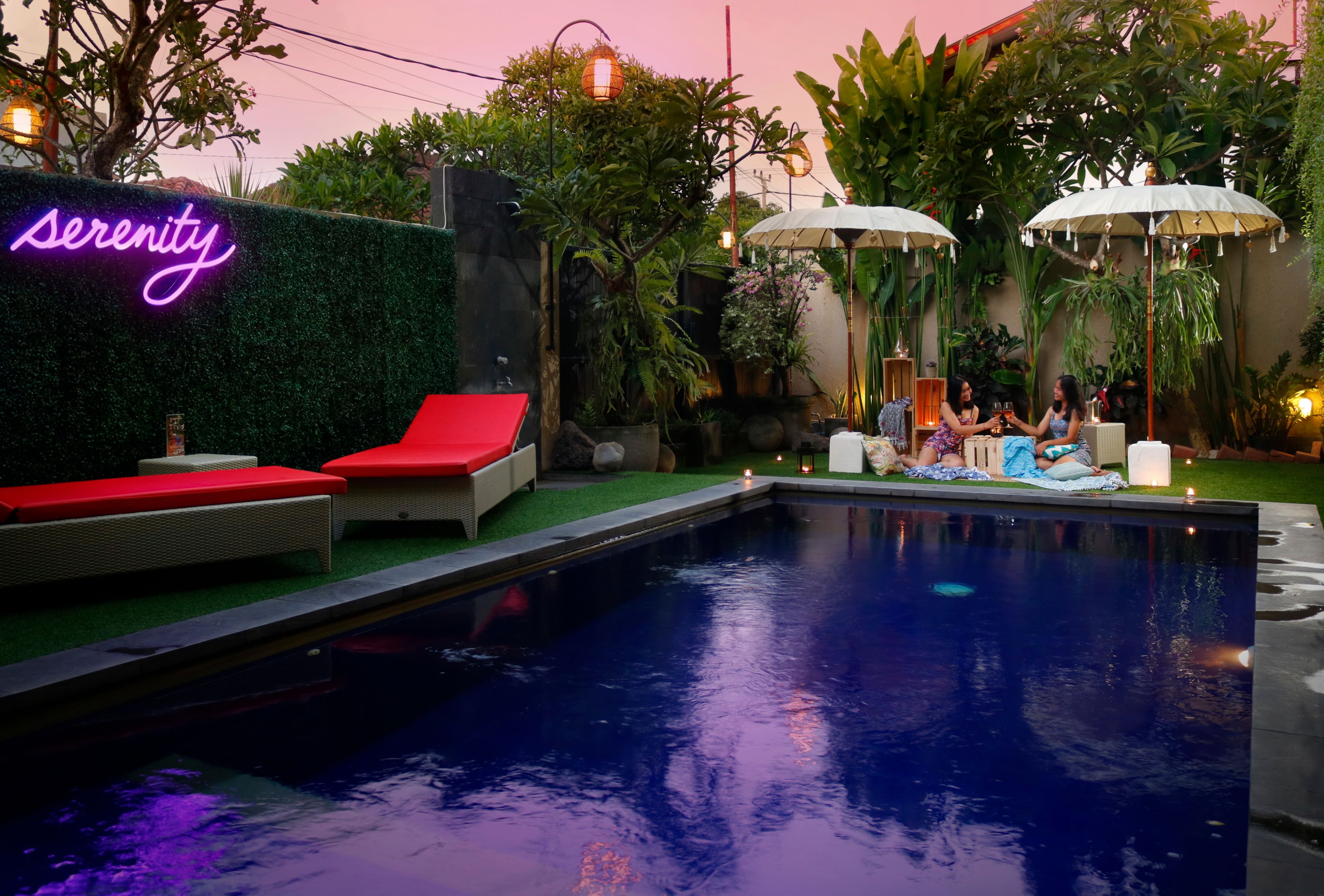 A pool area with red lounge chairs, a neon sign reading "serenity," and umbrellas.