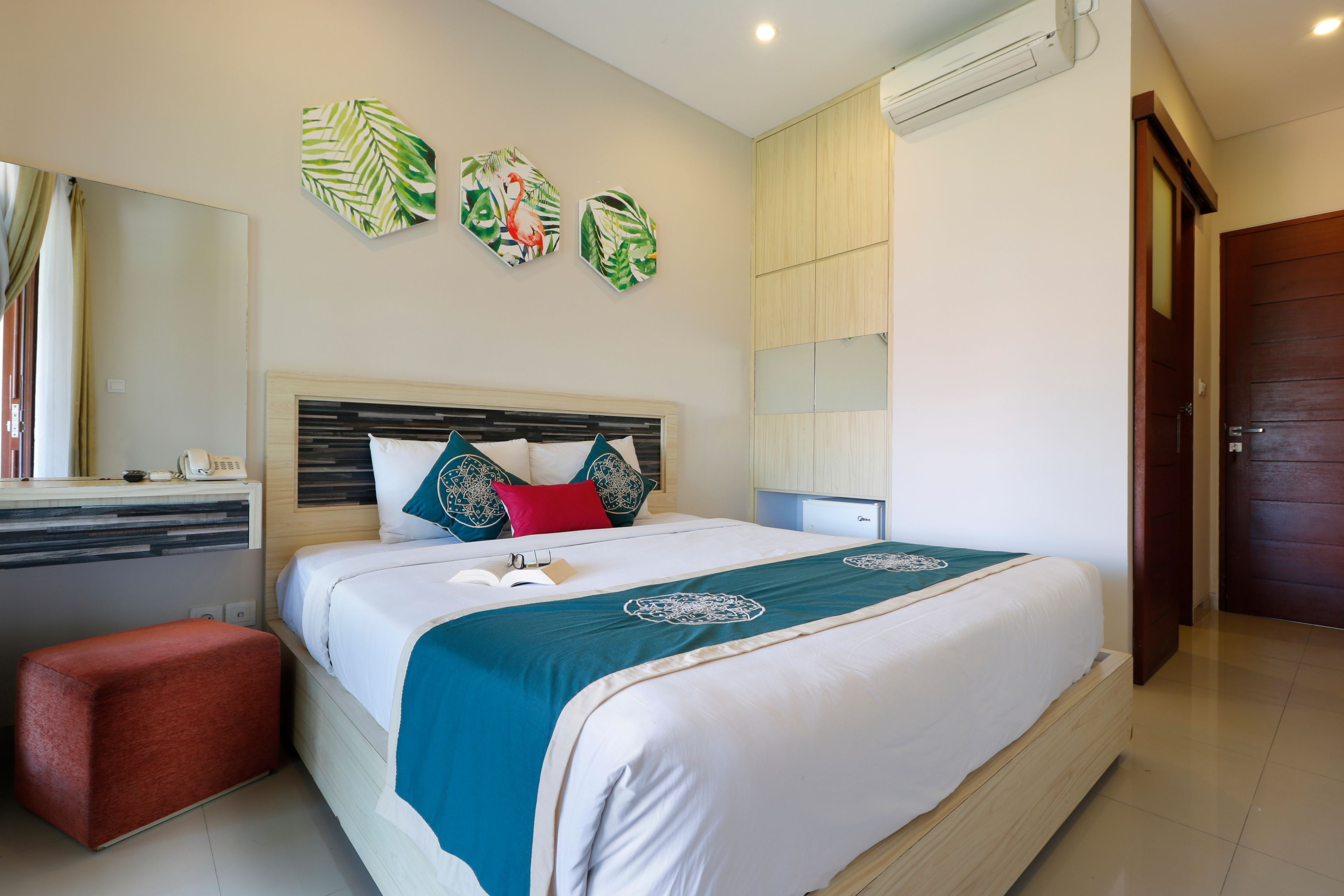 A neatly arranged bedroom with a bed, a bedside table, a red ottoman, and wall art.