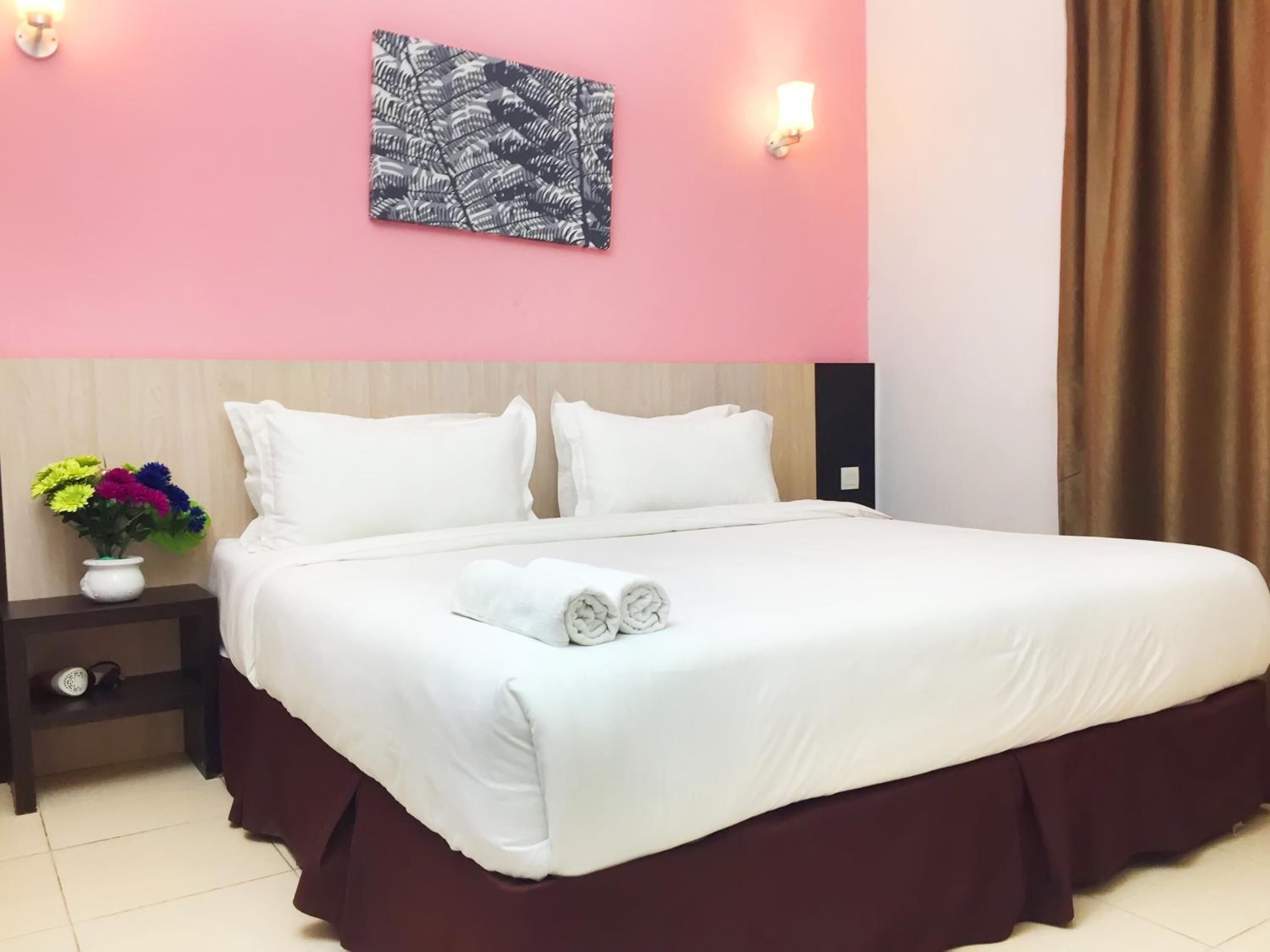 Habitación de hotel con pared rosa, cama con ropa de cama blanca, falda de cama marrón oscuro, mesita de noche con un jarrón de flores y una lámpara fijada a la pared.