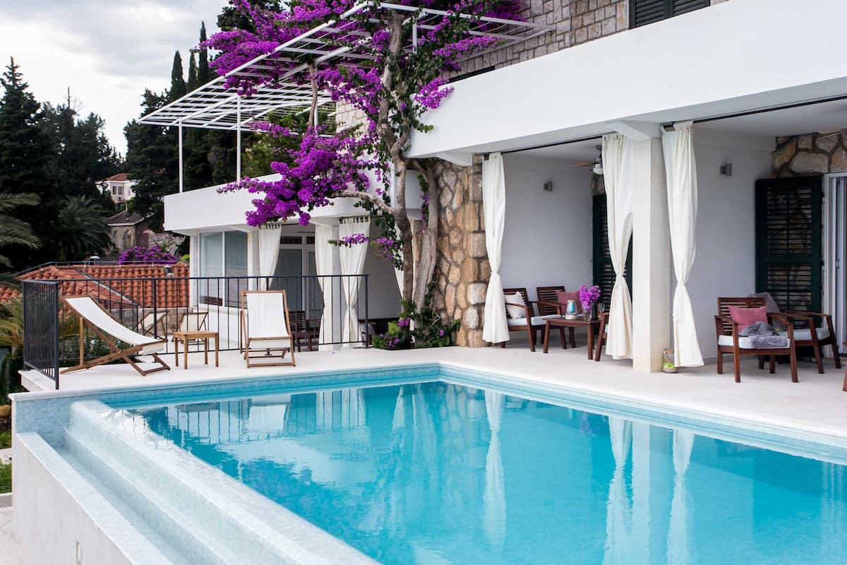 Une piscine dotée d’une pergola et de chaises, entourée d’un mur de pierre et d’un bâtiment blanc.