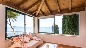 Ein Zimmer mit einem Bett, einem kleinen Tisch und großen Fenstern mit Blick auf das Meer und Bäume.
