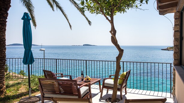 Eine Terrasse mit Holzmöbeln, einem Tisch mit einer Flasche und Obst, und Blick aufs Meer.