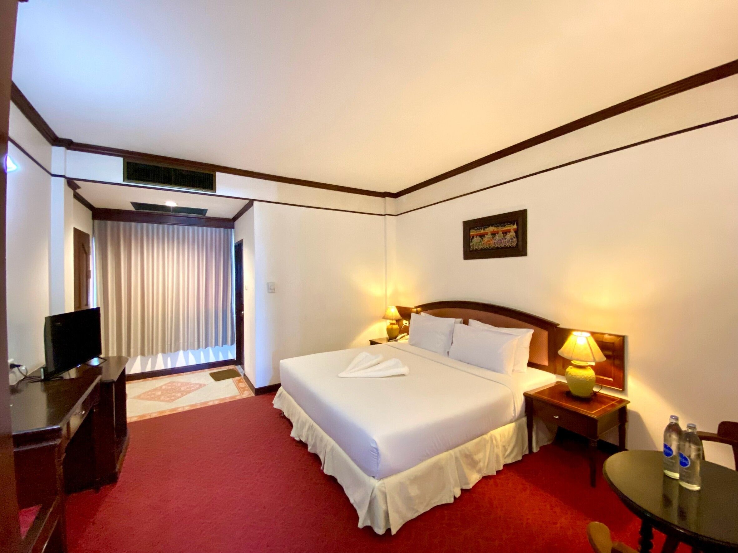 A hotel room with a bed, bedside lamps, a desk with a television, and a small table with water bottles.