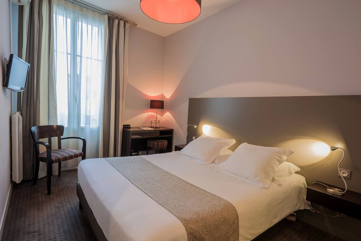 Une chambre d’hôtel avec un lit, un bureau, une chaise, une télévision et une fen être avec des rideaux.
